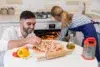 A couple prepares pizza in a bright kitchen, with fresh ingredients on the table and one person checking the oven.