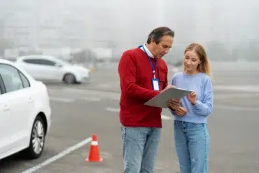 A man in a red sweater and a woman in a blue sweater discuss notes while standing beside a parked car in a foggy environment.