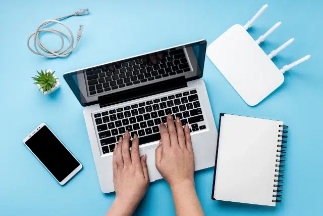 A woman typing on a laptop, with a phone and notebook beside her, focused on comparing internet service options.