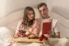 Couple in bed enjoying a cozy breakfast tray with wine, pastries, and roses while reading a book together.