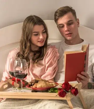 Couple in bed enjoying a cozy breakfast tray with wine, pastries, and roses while reading a book together.