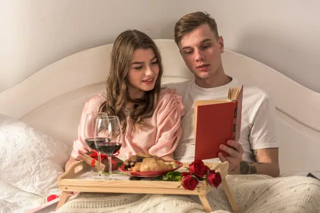 Couple in bed enjoying a cozy breakfast tray with wine, pastries, and roses while reading a book together.