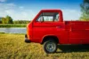 A bright red pickup truck parked on grassy land near a serene lake, with trees in the background under a clear blue sky.