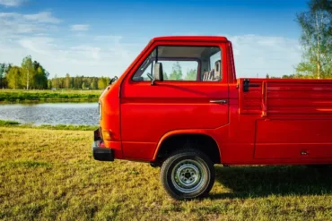 A bright red pickup truck parked on grassy land near a serene lake, with trees in the background under a clear blue sky.