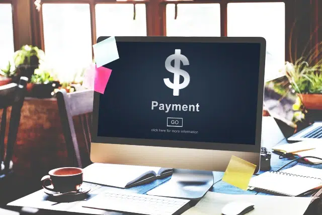 A desktop computer displaying a payment screen with a dollar sign, surrounded by a coffee cup and colorful sticky notes on a desk.