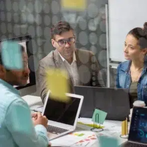 A team of professionals engaged in a meeting, surrounded by laptops and notes, with various documents and visuals on the table.