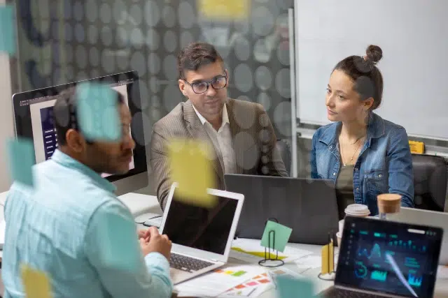 A team of professionals engaged in a meeting, surrounded by laptops and notes, with various documents and visuals on the table.