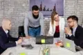 A group of four professionals collaborates around a table with laptops and charts, discussing strategies in a modern office setting.