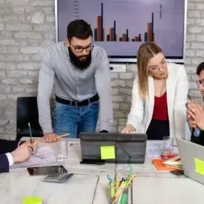 A group of four professionals collaborates around a table with laptops and charts, discussing strategies in a modern office setting.