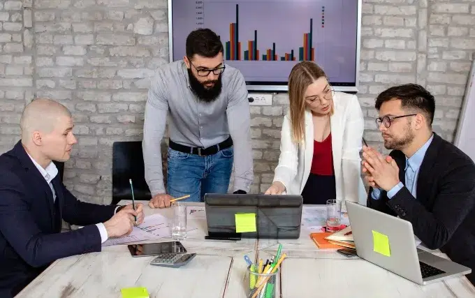 A group of four professionals collaborates around a table with laptops and charts, discussing strategies in a modern office setting.