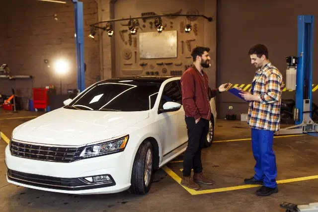A tech-savvy mechanic discusses diagnostics with a customer beside a modern white car in a repair shop.
