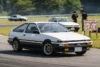 A white Toyota AE86 driving on a track, with a sign reading "For Sale" prominently displayed nearby..
