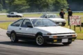 A white Toyota AE86 driving on a track, with a sign reading "For Sale" prominently displayed nearby..