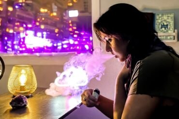 A woman is inhaling from an electronic cigarette while standing in front of a window.