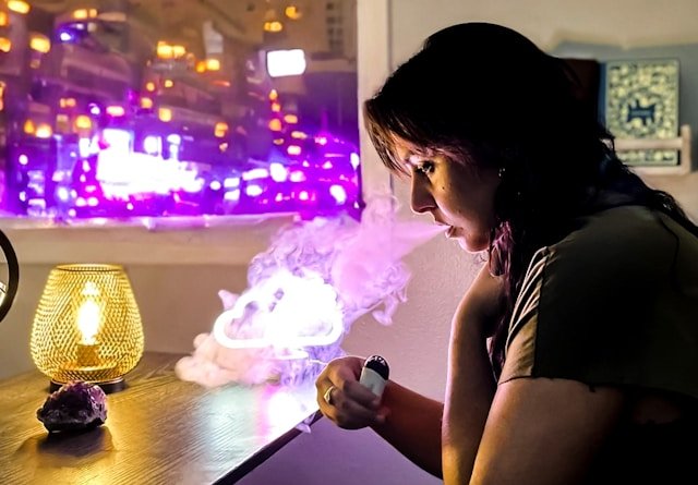 A woman is inhaling from an electronic cigarette while standing in front of a window.