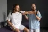 A physiotherapist assists a male patient with a shoulder exercise using a dumbbell in a therapy setting.