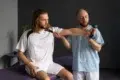 A physiotherapist assists a male patient with a shoulder exercise using a dumbbell in a therapy setting.