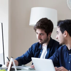 Two men collaborate at a desk, one using a laptop and the other working on a desktop computer, in a modern, bright workspace.