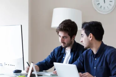 Two men collaborate at a desk, one using a laptop and the other working on a desktop computer, in a modern, bright workspace.