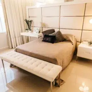 Sleek modern bedroom featuring a queen bed with beige bedding, white bench, and elegant lighting against a light-colored wall.