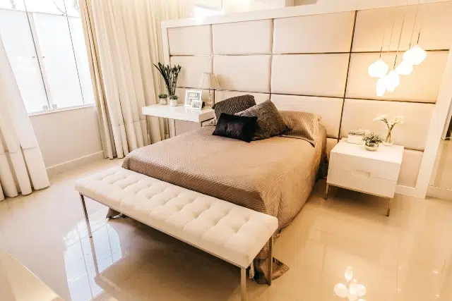 Sleek modern bedroom featuring a queen bed with beige bedding, white bench, and elegant lighting against a light-colored wall.