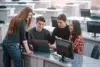 A group of four students collaborates around a laptop in a modern classroom setting, with computers and desks in the background.