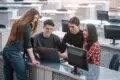 A group of four students collaborates around a laptop in a modern classroom setting, with computers and desks in the background.