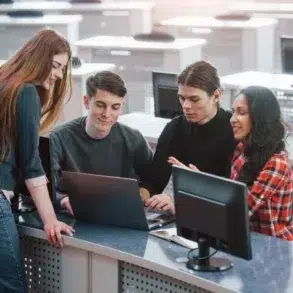 A group of four students collaborates around a laptop in a modern classroom setting, with computers and desks in the background.