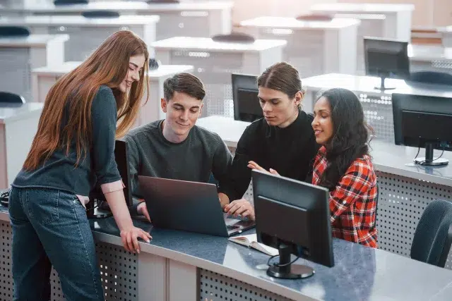 A group of four students collaborates around a laptop in a modern classroom setting, with computers and desks in the background.