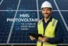 A worker in a hard hat and safety vest holds a tablet in front of solar panels, highlighting advancements in solar energy technology.