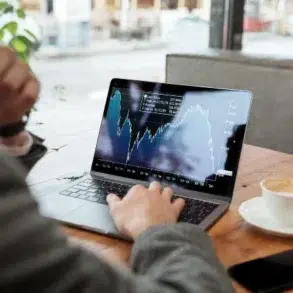 A man in a suit analyzes financial charts on a laptop at a cafe, with a cup of coffee and a smartphone beside him.