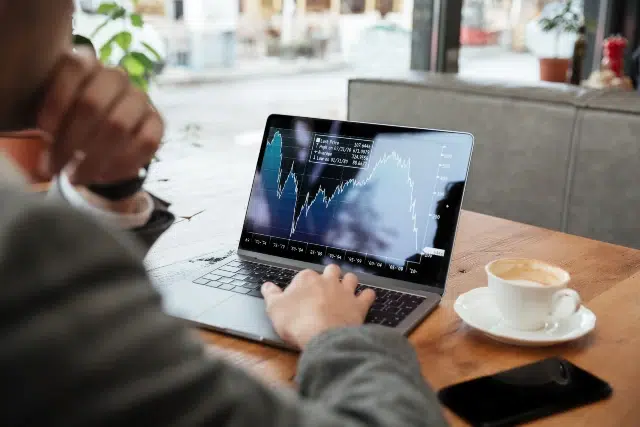 A man in a suit analyzes financial charts on a laptop at a cafe, with a cup of coffee and a smartphone beside him.