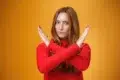 A woman in a red sweater gestures with crossed arms against a bright yellow background, conveying a message of rejection or defiance.