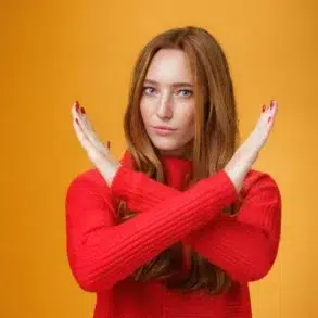 A woman in a red sweater gestures with crossed arms against a bright yellow background, conveying a message of rejection or defiance.