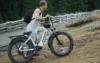 A person in a sleeveless shirt carries an electric bike along a rugged, grassy path near a wooden fence in a forested area.