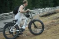 A person in a sleeveless shirt carries an electric bike along a rugged, grassy path near a wooden fence in a forested area.