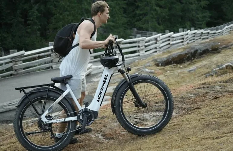 A person in a sleeveless shirt carries an electric bike along a rugged, grassy path near a wooden fence in a forested area.