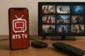 A smartphone displaying "RTS TV" on its screen, with a TV showing various stills and a remote control on a wooden surface.