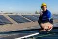 A worker in a hard hat and blue uniform crouches on a rooftop, holding a walkie-talkie near solar panels under a clear sky.