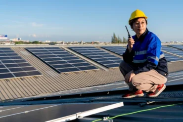 A worker in a hard hat and blue uniform crouches on a rooftop, holding a walkie-talkie near solar panels under a clear sky.