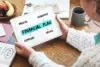 A person holds a tablet displaying a financial plan, outlining medical, insurance, expenses, and saving strategies on a wooden table.