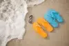 Colorful flip-flops in orange and blue beside sunglasses on sandy beach, with gentle waves lapping at the shore.