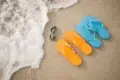 Colorful flip-flops in orange and blue beside sunglasses on sandy beach, with gentle waves lapping at the shore.