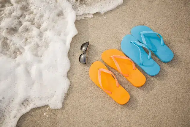 Colorful flip-flops in orange and blue beside sunglasses on sandy beach, with gentle waves lapping at the shore.