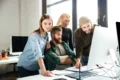 A group of four people gather around a computer, discussing ideas amidst a bright, modern office space.