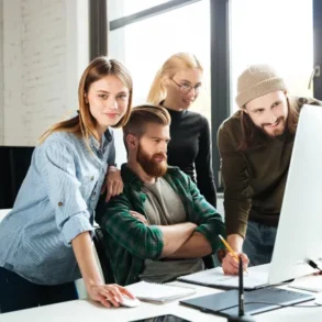 A group of four people gather around a computer, discussing ideas amidst a bright, modern office space.