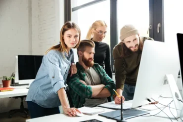 A group of four people gather around a computer, discussing ideas amidst a bright, modern office space.