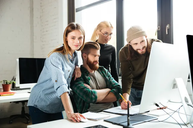 A group of four people gather around a computer, discussing ideas amidst a bright, modern office space.