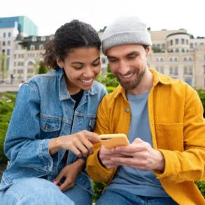 A young man and woman sit outdoors, closely examining a smartphone together, with greenery and urban buildings in the background.