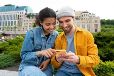 A young man and woman sit outdoors, closely examining a smartphone together, with greenery and urban buildings in the background.